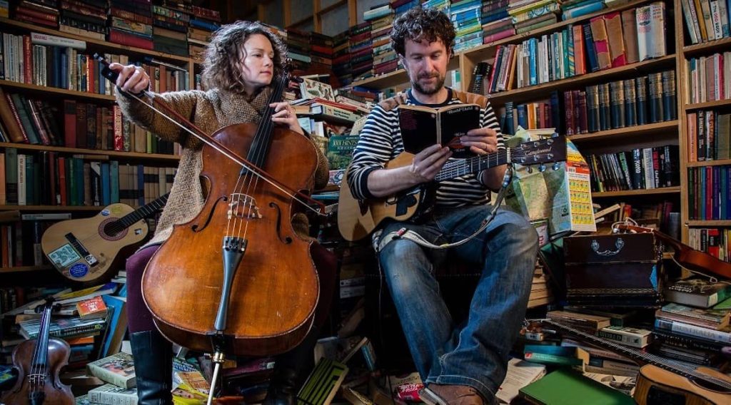 Beth and ben sit amongst bookshelves and piles of books. Beth is ready to play the cello she holds; Ben has a guitar tucked under his arm but an open book in his hands, that Beth leans over to look at.