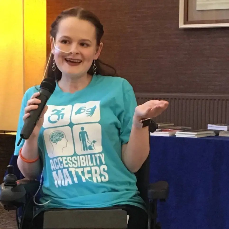 Hannah photographed as she talks, with a microphone in one hand and her other hand held out. She is sitting in a power chair and has a feeding tube. She wears a turquoise accessibility matters t-shirt and has dark red hair in french plaits.