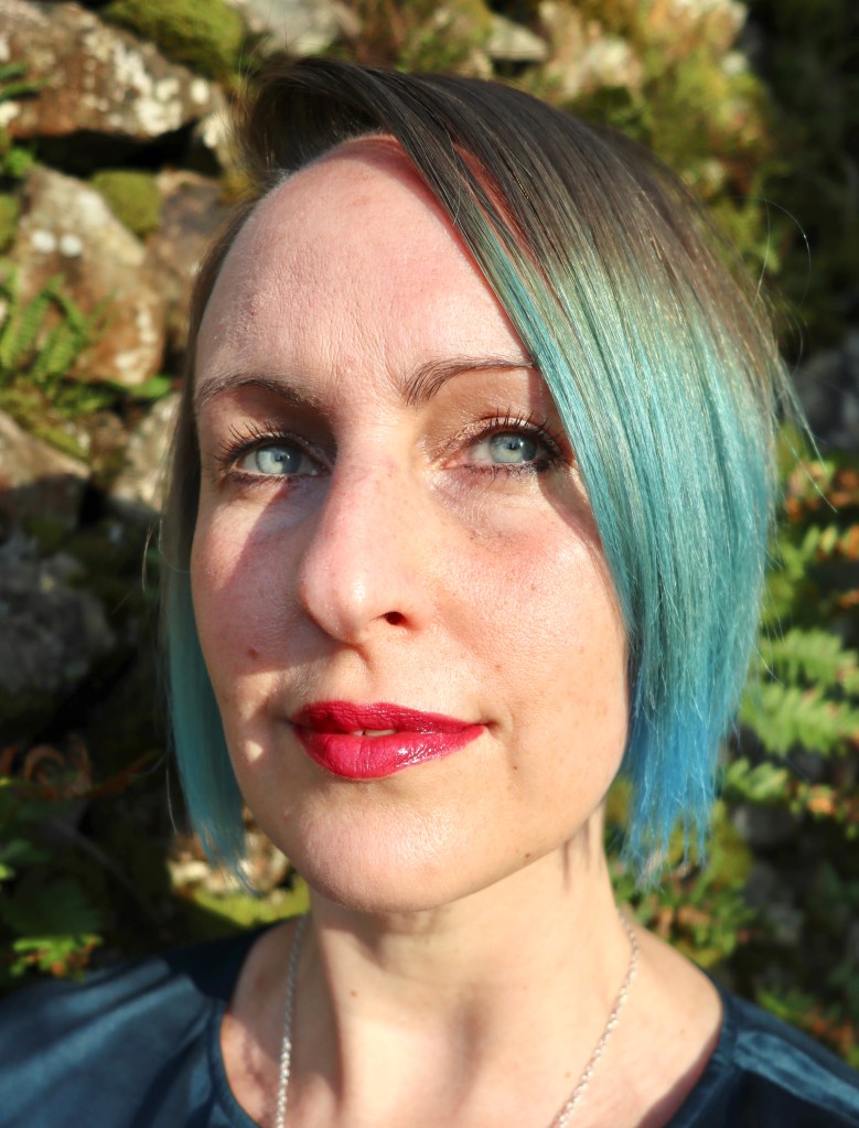 Polly's head in the sun in front of a mossy dry stone wall. She has blue eyes and brown and blue hair in a short bob.